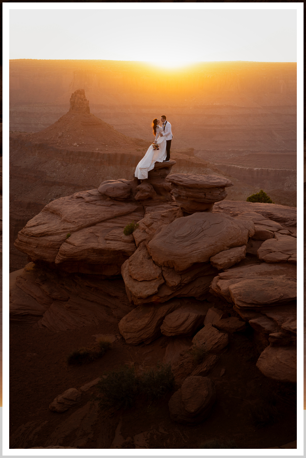 A wedding couple in Dead Horse State Park standing on a large stack of deep red stone at sunset with cinematic hazy lighting near the edge of the cliff.